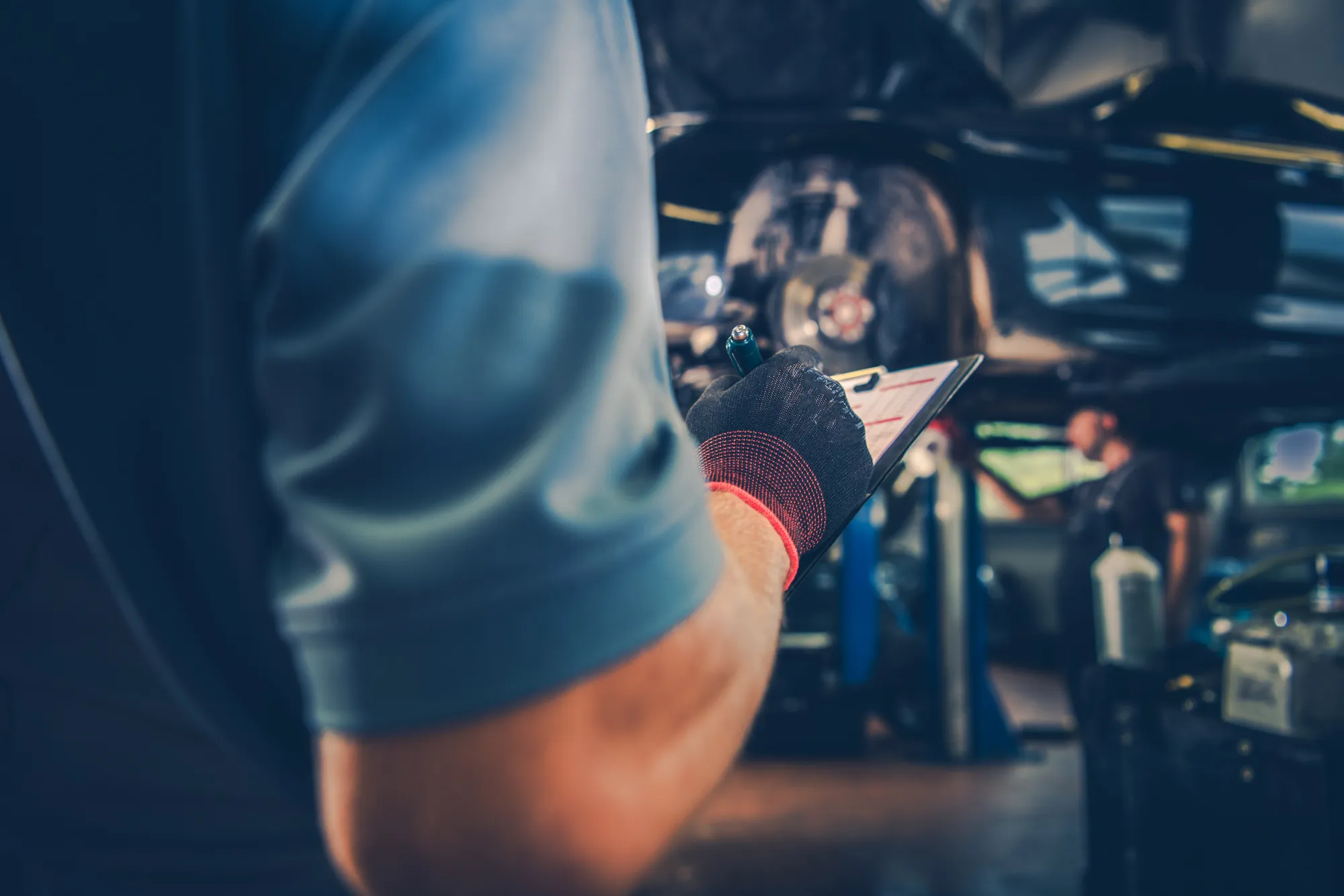 A mechanic in a work glove holds a clipboard and pen while inspecting a car raised on a lift in a repair shop.